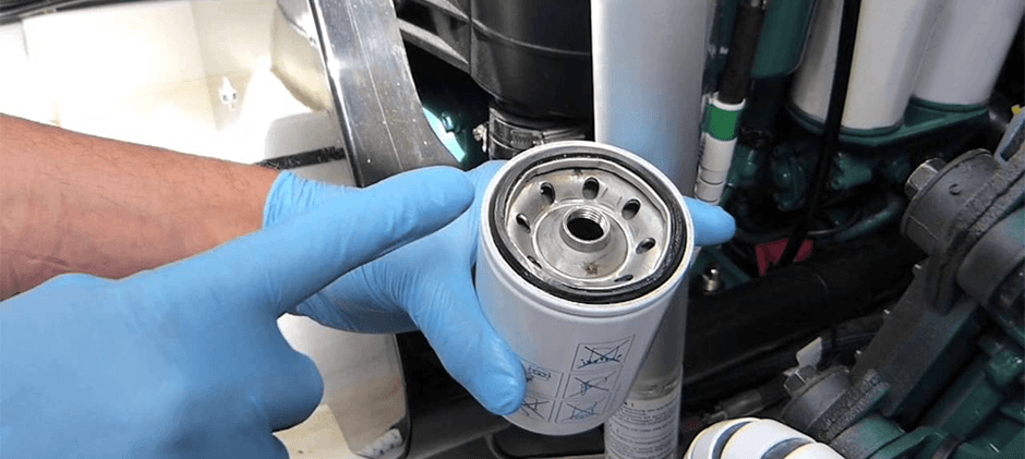 A person wearing gloves performs a DIY fuel filter replacement on a heavy equipment engine, holding a new aftermarket filter.