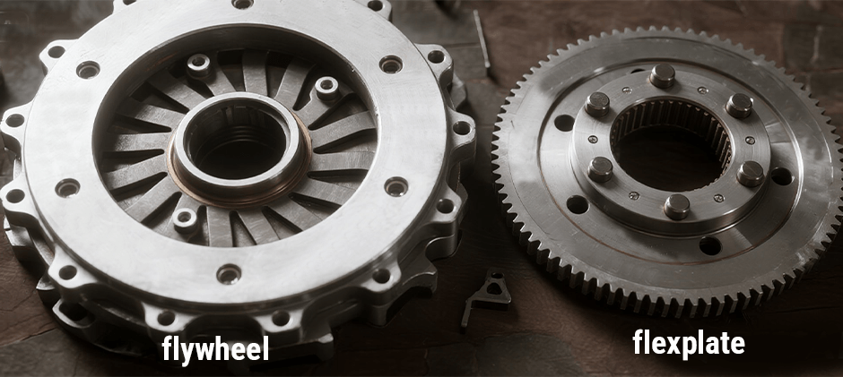 A side-by-side comparison showing a heavy, thick flywheel on the left and a thin, lightweight flexplate with a large ring gear on the right. The image is labeled to clearly identify each part and demonstrate their physical differences.
