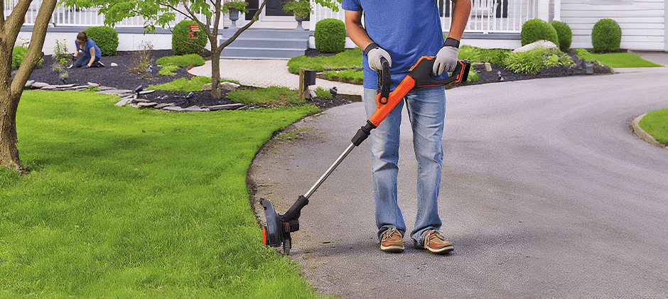 A person creating a sharp, clean edge along a paved driveway using a battery-powered string trimmer.