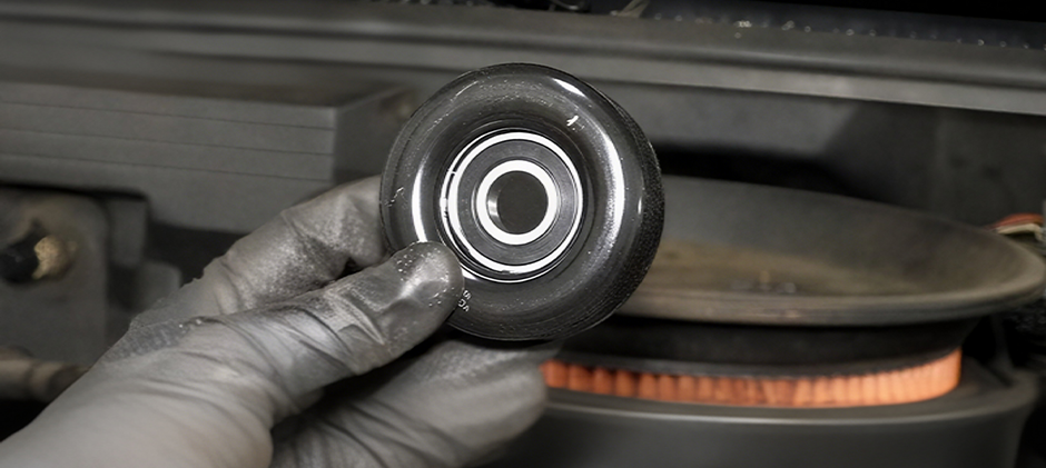 A mechanic's gloved hand holding a standalone idler pulley, showing the central bearing up close in front of an engine.