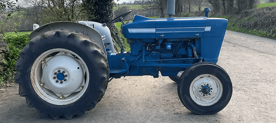 Side profile of a blue and grey Ford 2000 utility tractor, highlighting its engine and classic design, parked on the side of a country road.