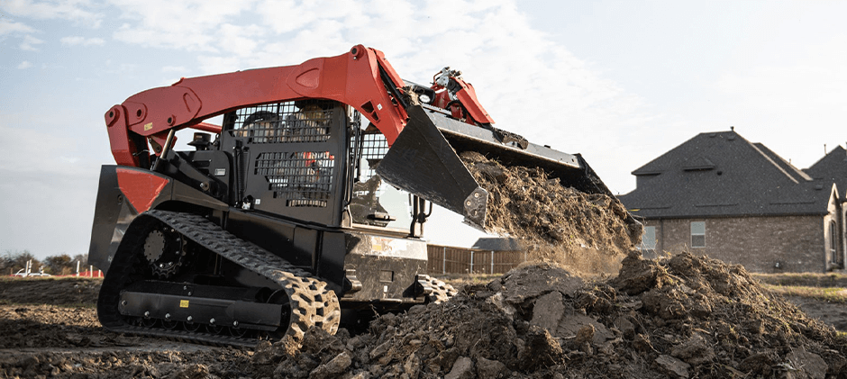  A powerful compact track loader dumping a large bucket of dirt and rocks on a construction site.