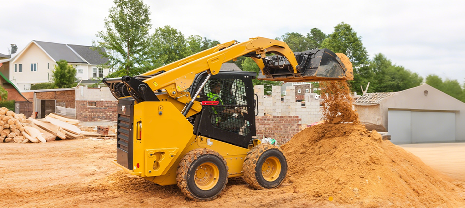 A yellow Caterpillar skid steer loader in action at a construction site, lifting its bucket to dump a load of dirt onto a pile.