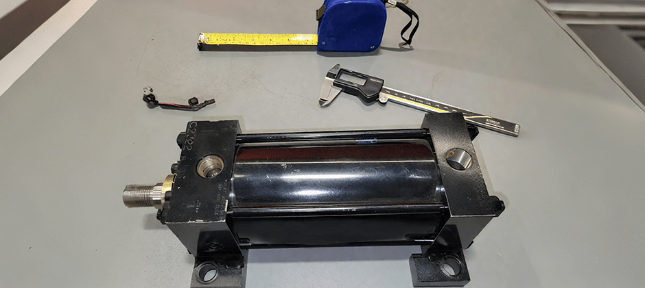  A black tie-rod hydraulic cylinder on a workbench with the essential tools for measurement: a digital caliper and a tape measure.