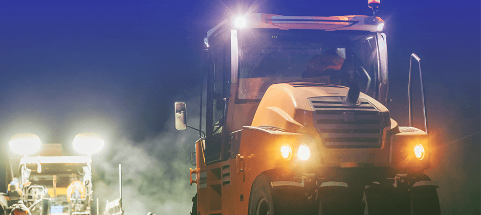 Two pieces of heavy equipment working at dusk; one in the foreground has its headlights on, while the machine in the background uses powerful work lights to illuminate the area.
