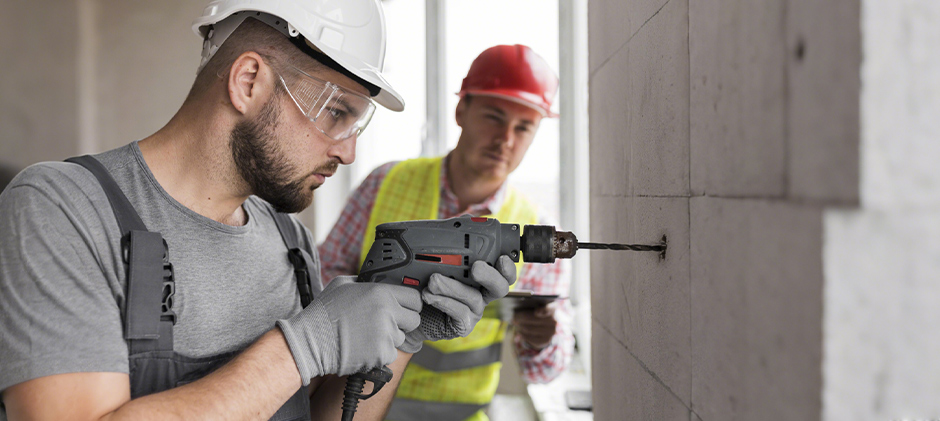 A construction worker in a hard hat using a hammer drill to make a hole in a concrete wall.