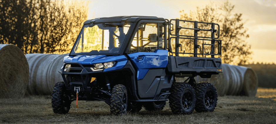 A blue Can-Am Defender 6x6 UTV with a long cargo bed parked in a field with large hay bales.