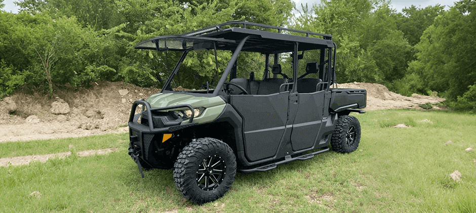 A green Can-Am Defender MAX 6-Seat Models with a roof rack and flip-up windshield parked in a grassy area.