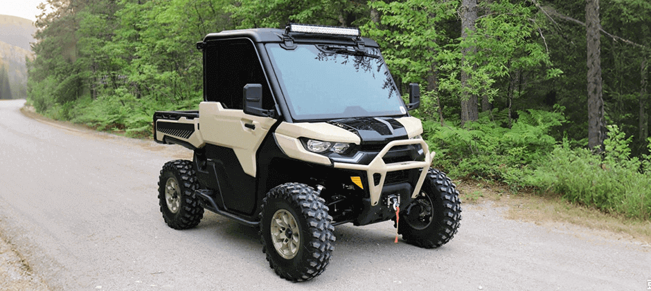 A tan Can-Am Defender HD10 UTV equipped with a winch and light bar parked on a wide dirt trail in a forest.