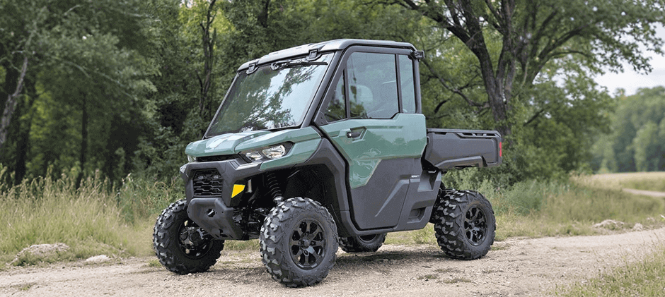A dark green Can-Am Defender HD9 utility side-by-side on a narrow path surrounded by lush green trees.