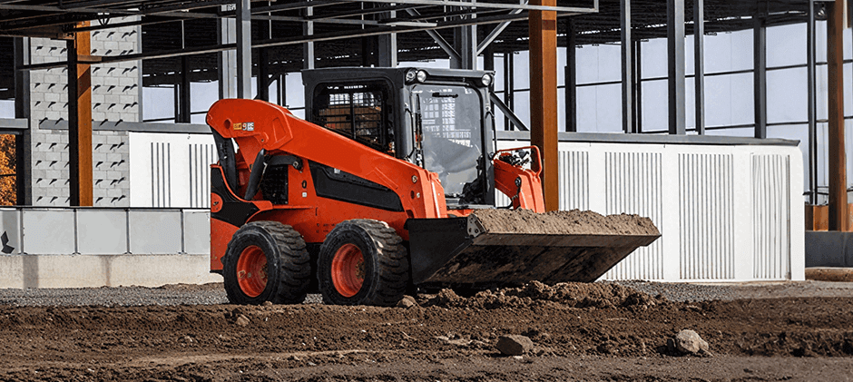 An orange Kubota skid steer loader moves dirt with its bucket at a construction site.