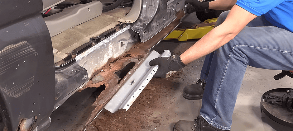 A mechanic holding a new replacement rocker panel next to a car with a completely rusted-out rocker panel, showing the effects of corrosion.