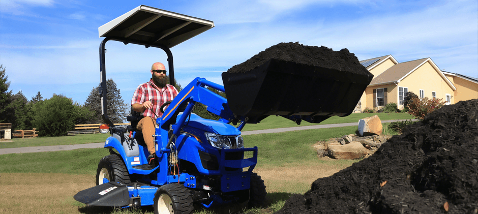 An operator on a New Holland Workmaster 25S compact tractor with a sun canopy, lifting a full loader bucket of mulch for a landscaping project.