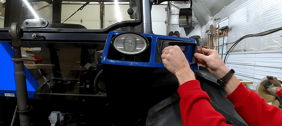 A person's hands performing a DIY repair on the light housing of a blue tractor in a workshop.