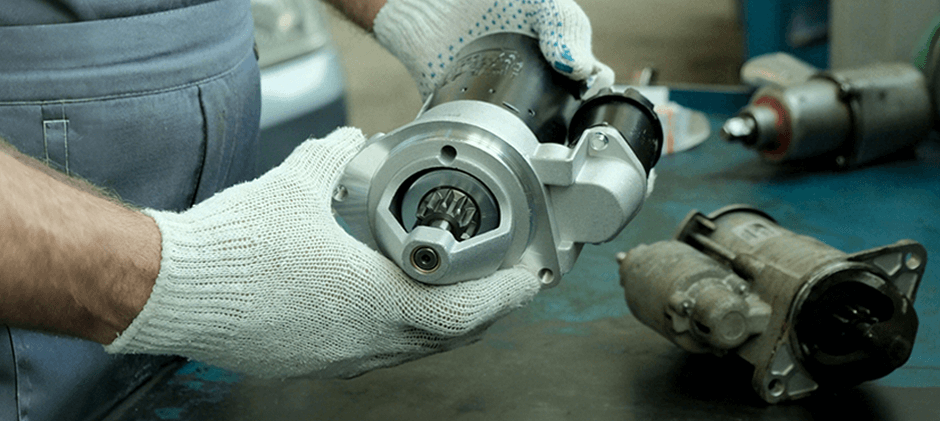 A mechanic wearing gloves holds a shiny new starter motor, with the old, dirty starter motor visible in the background for comparison.