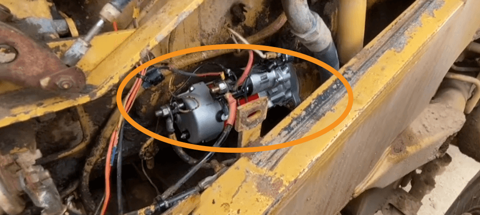 A starter motor installed in the engine bay of a piece of heavy machinery, with an orange circle highlighting its exact location on the engine block.