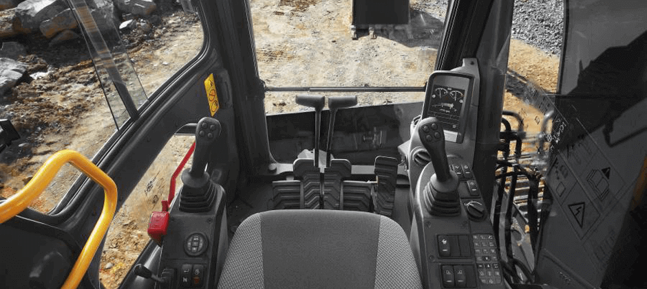 View from inside an excavator cab, showing the operator's seat, two joysticks, foot pedals, and the view of the work site.