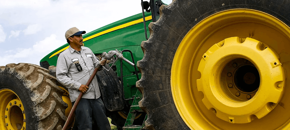 A farmer holding a fuel nozzle next to the large yellow wheel of a green tractor.