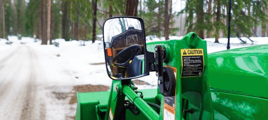 A rear view mirror mounted on a green loader, reflecting the operator's area and a barn on a snowy day, demonstrating the need for clear visibility.