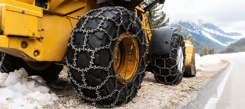 Close-up of heavy-duty snow chains installed on the large tire of a yellow wheel loader on a snowy mountain road.