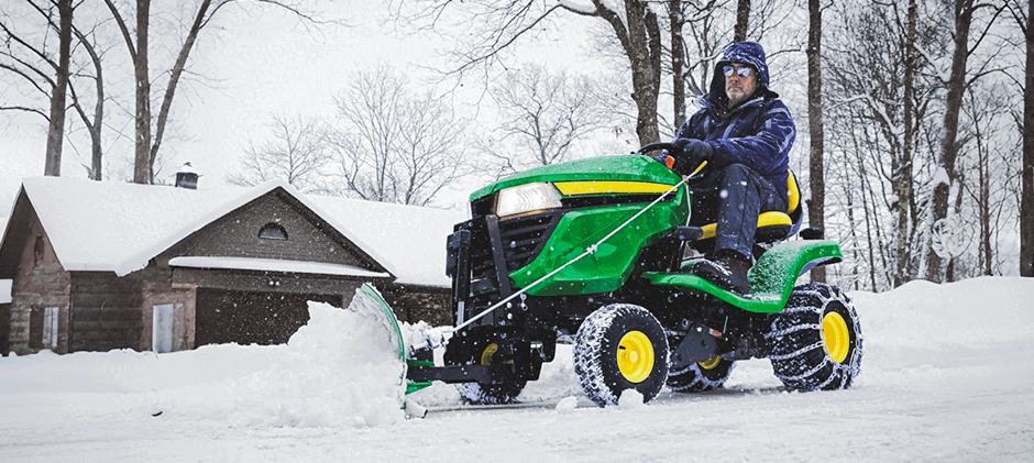 A man operating a John Deere X300 tractor with a yellow snow blade attachment to clear deep snow from a residential driveway in winter.