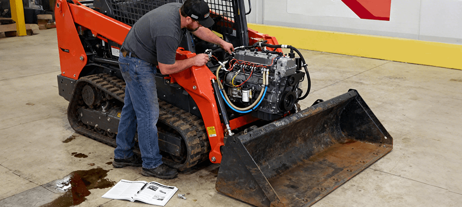 A mechanic performs maintenance on a Kubota V2403 diesel engine inside a compact track loader, with a service manual open on the floor.