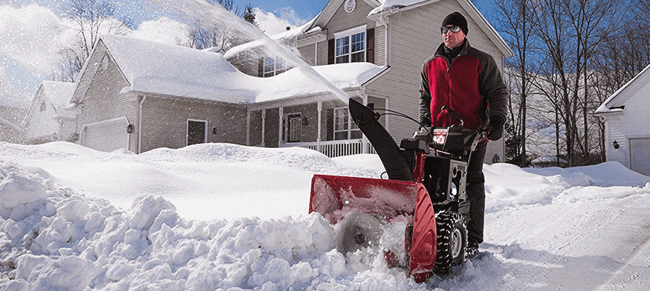 A man in winter gear clears his snowy suburban driveway with a red snow blower on a sunny day, with snow-covered houses in the background.