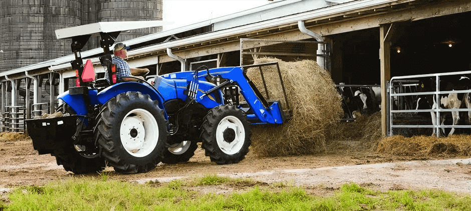 A blue New Holland Workmaster 50 tractor using its front-end loader to move a large round hay bale on a farm.