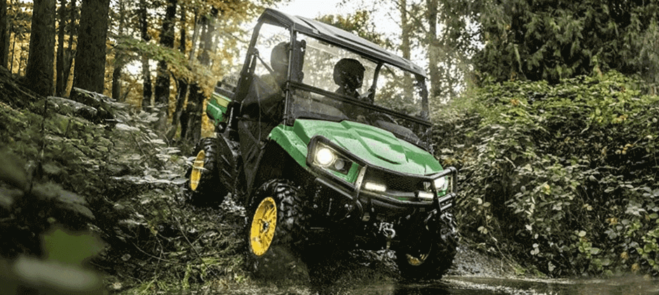A John Deere Gator 4x4 utility vehicle climbing a steep and muddy hill in a dense forest.