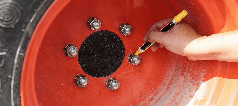 A close-up of a hand using a yellow torque seal marker to draw a line on a lug nut on a red tractor wheel.