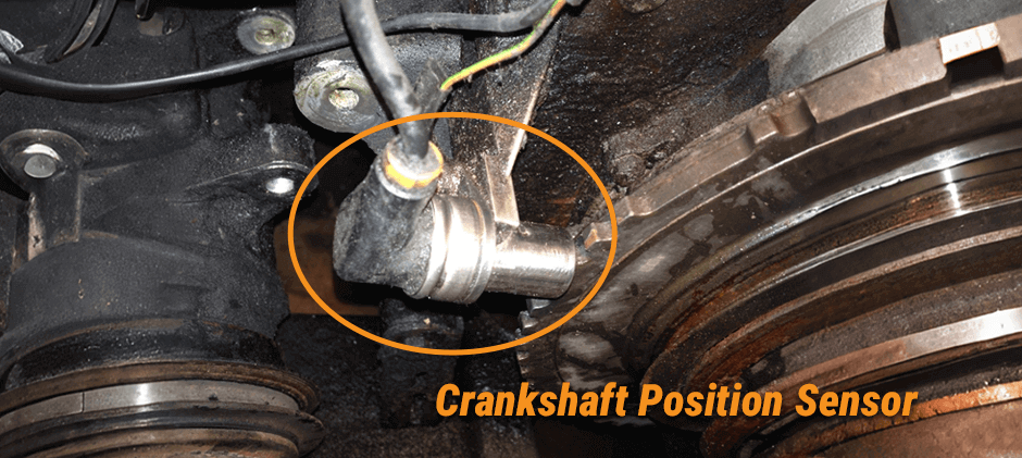 A close-up photo of a crankshaft position sensor installed in a heavy equipment engine block. The sensor is circled in orange to clearly show its location near the crankshaft.