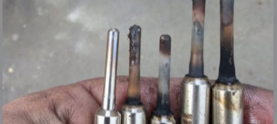 A mechanic's hand holding five glow plugs, showing a new plug next to four used ones with carbon buildup, a broken tip, and heat discoloration.