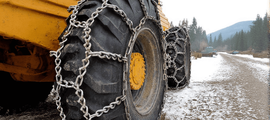 Close-up of heavy-duty snow chains installed on the large tire of a piece of yellow construction machinery in the snow.