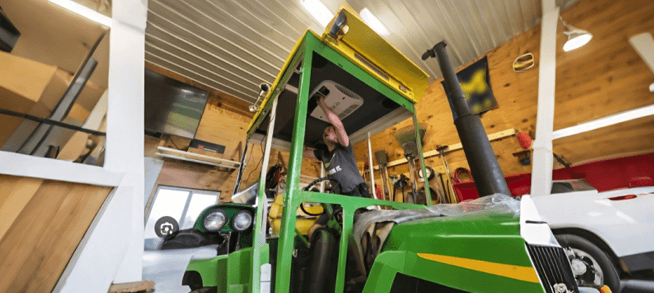 A technician working on a roof-mounted HVAC unit inside a green tractor cab to access the blower motor.