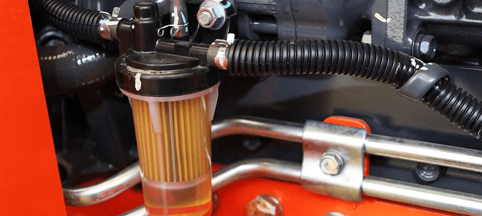 A close-up of a diesel fuel filter with a clear water separator bowl installed in a heavy machinery engine bay, showing the fuel lines and internal filter element.