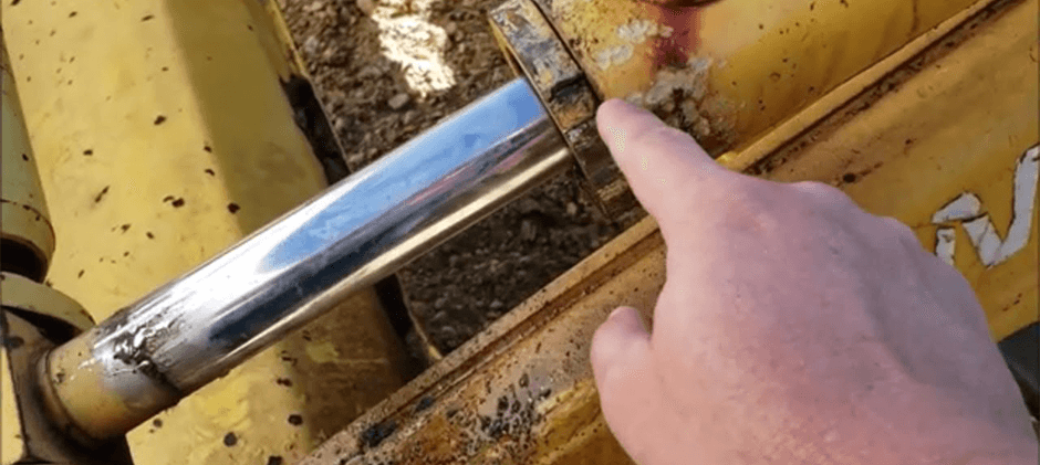 A mechanic's hand pointing to the end cap, or gland, of a hydraulic cylinder on a piece of heavy machinery.