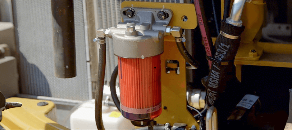 A primary fuel filter and water separator with a clear bowl mounted on the side of a dirty heavy equipment engine.