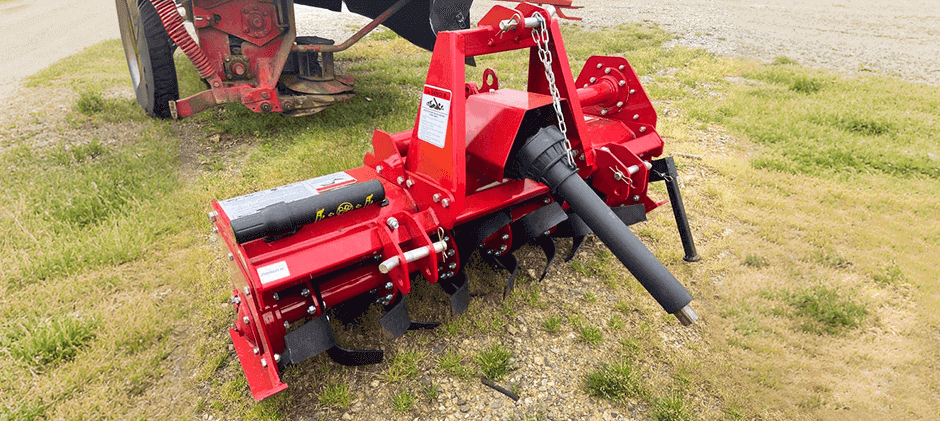 A red PTO-driven rotary tiller attachment for a tractor, showing the tines and the driveshaft.