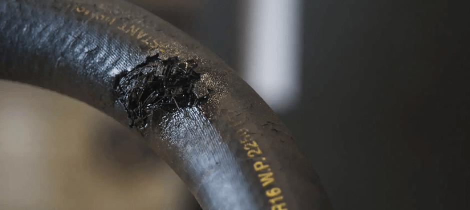 An extreme close-up of a burst hydraulic hose with frayed internal reinforcement wires visible through the damaged outer cover.