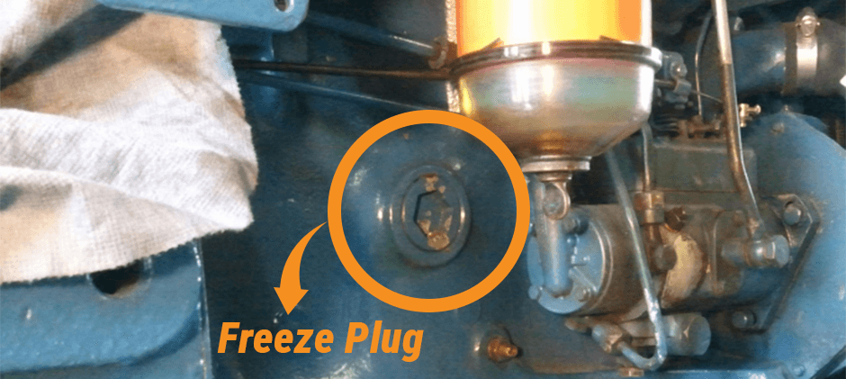 A detailed view of a blue engine block with an orange circle and arrow pointing directly to a freeze plug, helping to identify its location and appearance.
