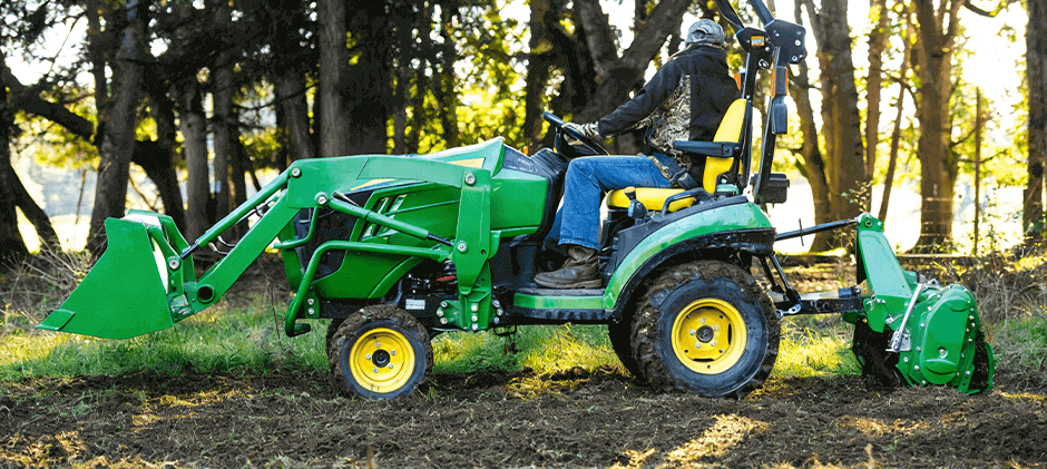 A person operating best john deere tractor for small farm with a front loader and a rear tiller attachment to prepare a garden bed.