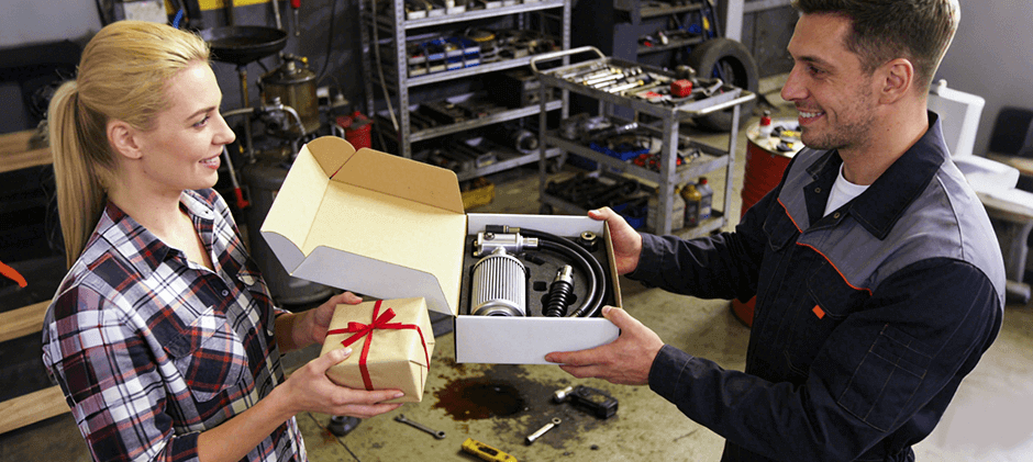 Woman giving a gift box containing hydraulic parts and filters to a mechanic in a workshop, representing practical Valentine’s Day gift ideas for heavy equipment professionals.