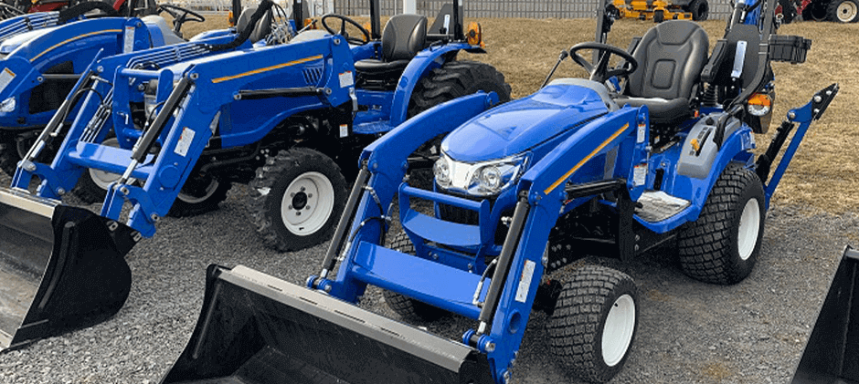 Several new blue New Holland Workmaster 25S tractors with front loaders parked in a row on a gravel lot.