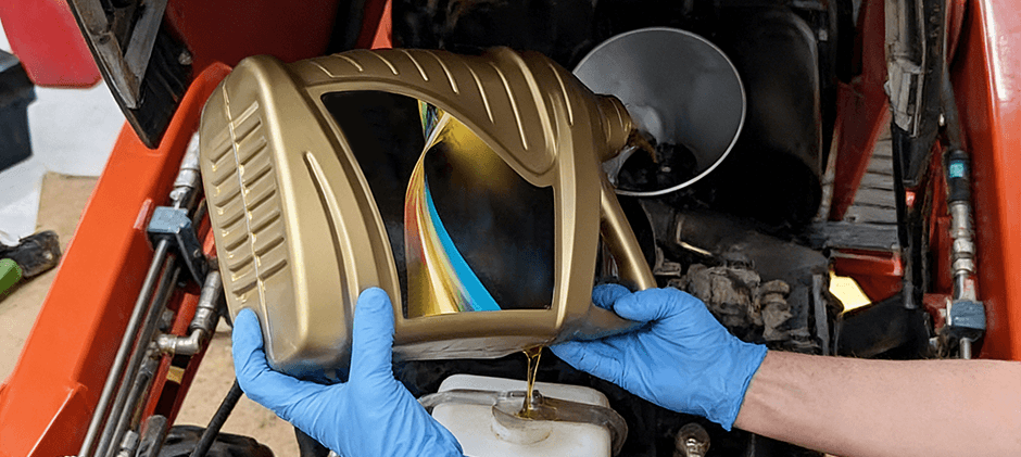 A close-up of a mechanic in blue gloves carefully pouring oil from a gold jug into an orange machine's engine.