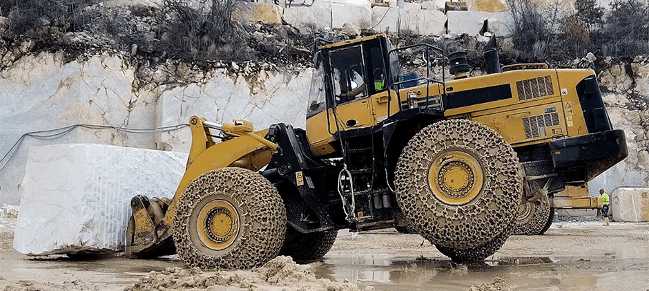 A large yellow wheel loader with tire chains lifting a massive block of white stone in a quarry, with its rear wheels slightly off the ground.