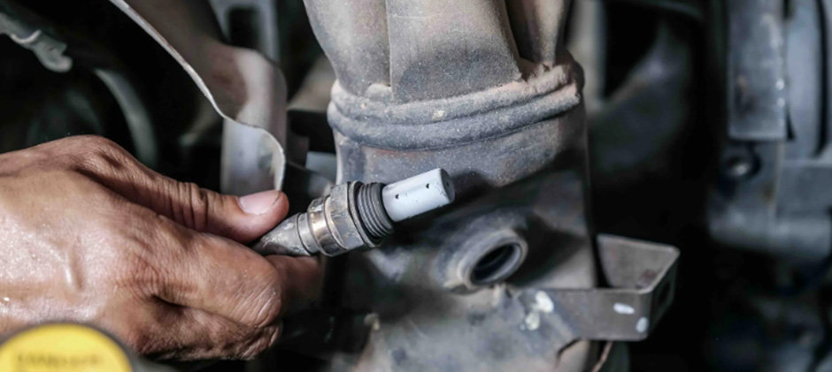 A mechanic's hand unscrewing a downstream oxygen sensor from the exhaust manifold of a heavy-duty engine.