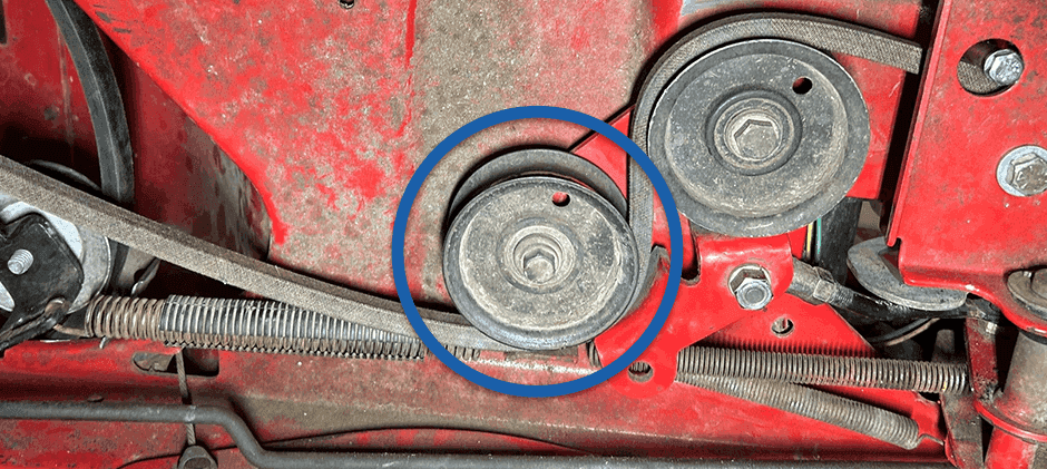 A close-up of a worn and dirty drive belt tensioner pulley on a red mower deck, circled to show a common point of failure.