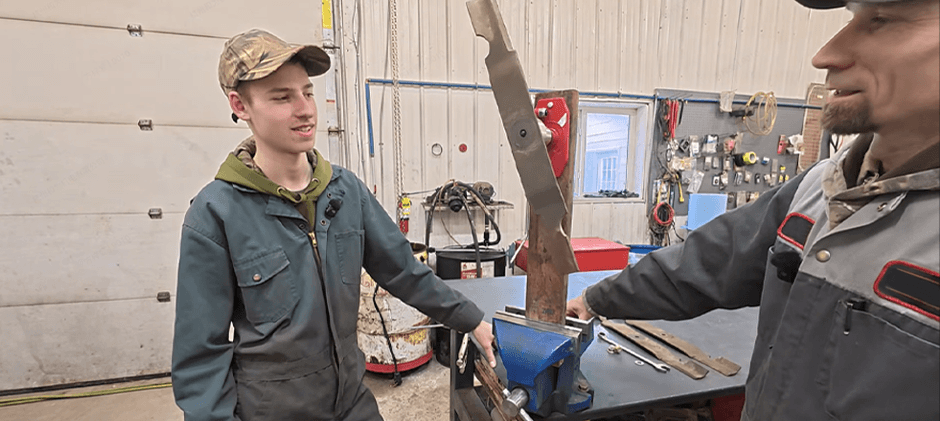 Two mechanics in a workshop, one younger and one older, inspecting a large mower blade clamped in a vise before sharpening and balancing.