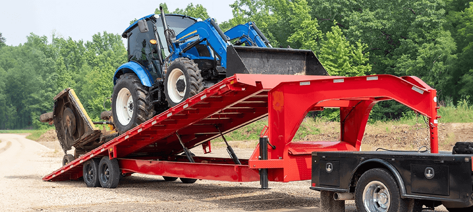 A blue tractor with a front loader being driven onto a red tilt-deck trailer that is angled down to the ground.