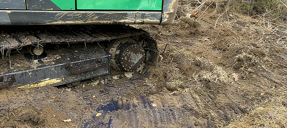 A close-up of a muddy track on a compact loader showing a visible puddle of leaked hydraulic or steering fluid on the ground beneath it.